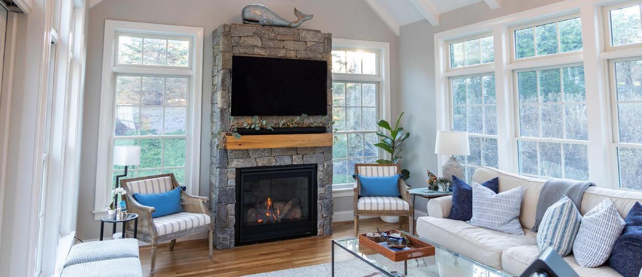 A living room with large bay windows and a statement stone fireplace with television and wooden mantle